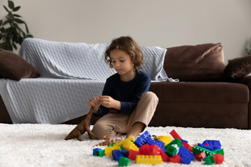 Little boy playing with toy dinosaur and colorful plastic blocks alone, sitting on warm floor carpet at home, adorable child kid engaged in educational activity, spending leisure time in living room