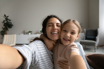 Head shot portrait of smiling mother and daughter taking selfie, hugging and looking at camera,...