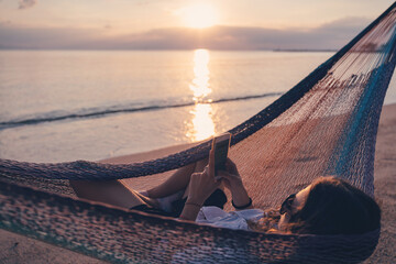 Young woman in a hammock with a smartphone in her hands on the shore of a tropical beach at sunset