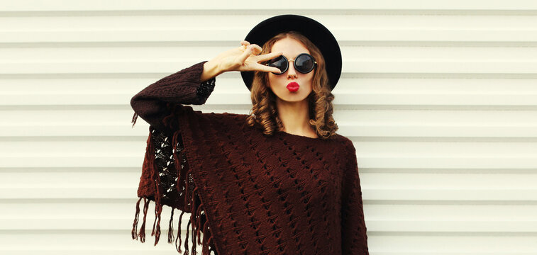 Portrait of beautiful young woman blowing her lips sending sweet air kiss wearing a brown knitted poncho, black round hat on white background