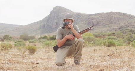 Kneeling caucasian male survivalist holding hunting rifle, looking using binoculars in wilderness - Powered by Adobe