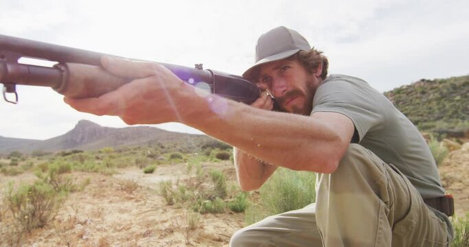 Kneeling caucasian male survivalist taking aim with hunting rifle in wilderness