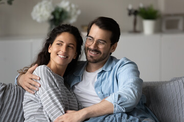 Head shot of dreamy young couple family hugging, looking in distance, sitting on cozy couch at home, happy beautiful woman and man visualizing good future together, dreaming of new opportunities