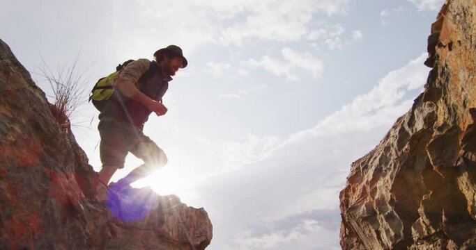Bearded caucasian male survivalist with backpack jumping across rocky mountain ravine in wilderness