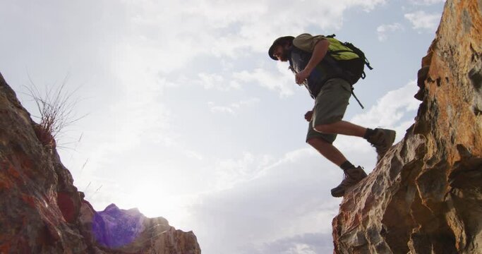 Bearded caucasian male survivalist with backpack jumping across rocky mountain ravine in wilderness