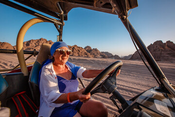Happy elderly woman in a blue headscarf driving a buggy in the Egyptian desert with mountains in the background.