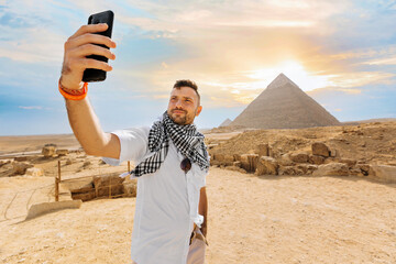 A young man, in front of the pyramid, raises his phone, takes a selfie with his fingers in front of...