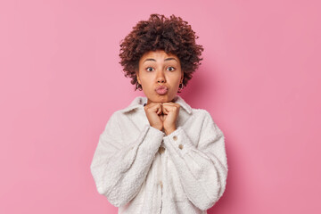 Romantic beautiful woman with curly hair keeps lips folded hands under chin looks with adoration at something going to kiss you wears fashionable white fur jacket isolated over pink background