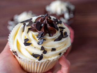 close-up in the hand of a cupcake with white cream decorated with pieces of dark chocolate. Sweets, delicious dessert. Calorie content, unhealthy food