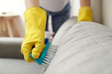 Woman cleaning grey sofa with brush and detergent at home, closeup