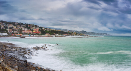 Fototapeta na wymiar Waving sea over coast line and water bay in winter, Riviera dei Fiori, Liguria, Italy. Sanremo town in the background. Dramatic stormy sky at sunset. Long exposure.