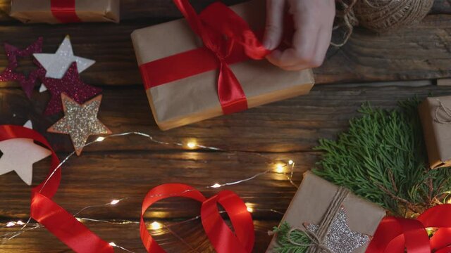 female woman hands wrapping Christmas presents gift box with brown craft paper, tying red ribbon bow on wooden desk with new year ornaments: stars light garland and green coniferous. top view DIY home