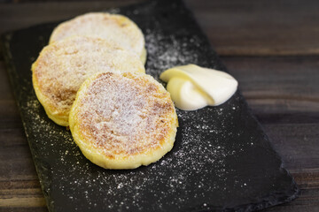 pancakes with cottage cheese with sour cream on a black board and a black background , sprinkled with powdered sugar