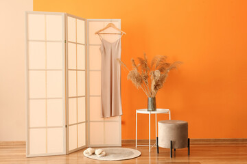 Pampas grass in vase on table and folding screen with dress near orange wall