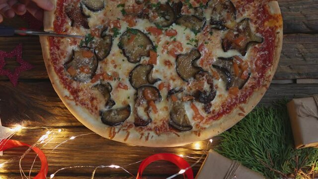 Cutting Vegetarian Pizza With Sliced Eggplants Using Scissors On Wooden Table Decorated With Red Ribbon And Golden Lights Garland And Green Branch Of Thuja. Life Hack New Way To Cut Hot Pizza 