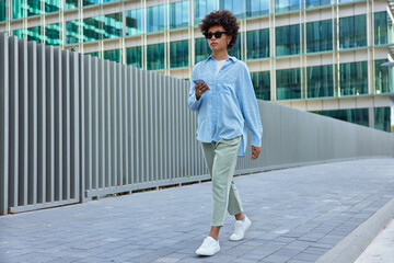 Pretty stylish hipster girl walks with smartphone in city being on her way to work browses cellular makes publication for her blog chats and sends messages poses against modern glass building