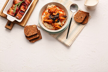 Bowl with delicious oatmeal and baking dish with prunes baked in crispy bacon on light table