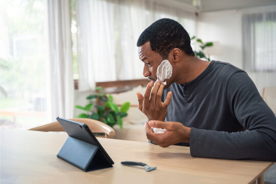 African American man applying cream on his face and using digital tablet pretend to be mirror before shaving his beard - Powered by Adobe