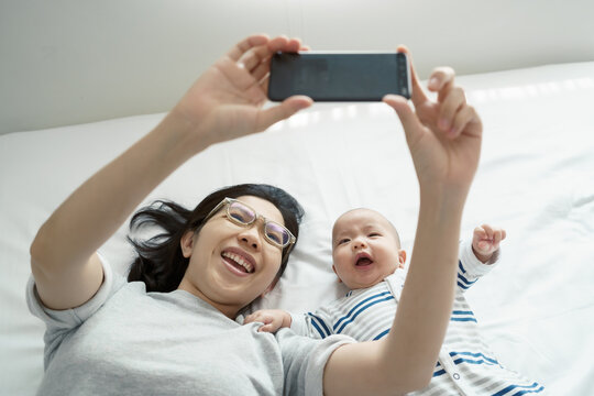 Happy Smiling Asian Young mother and her little cute baby boy lying on bed taking selfie photos with smartphone together,