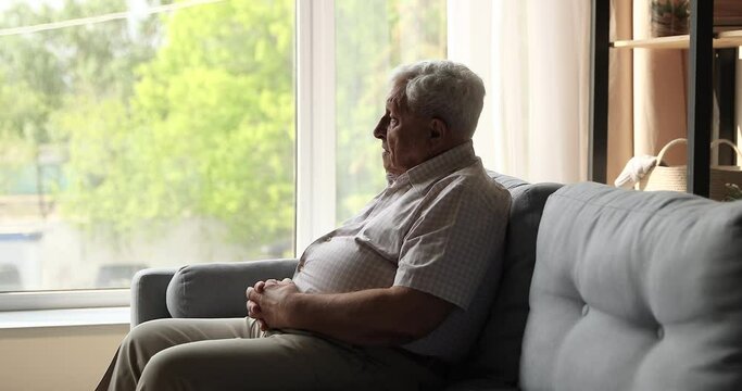 Sad unhappy mature male retiree sit on couch alone look at window with spring view feel abandoned depressed suffer of weakness ageing retirement problems. Upset elderly man grieving about passed youth