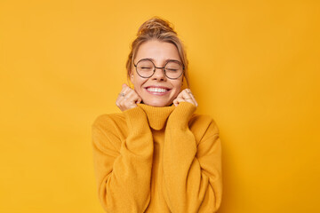 Cheerful young woman enjoys pleasant memories wears soft comfortable jumper and round spectacles keeps eyes closed smiles pleasantly isolated over yellow background recalls nice moment in life