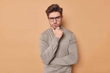 Fototapeta premium Waist up shot of serious man holds chin looks attentively at camera has self confident expression wears transparent glasses and turtleneck isolated over beige background considers something.