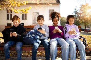 Group of multicultural children sitting on bench and using their smart phones surfing internet -...
