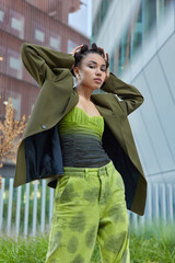 Outdoor shot of fashionable young woman stands in self confident pose keeps hands behind head...
