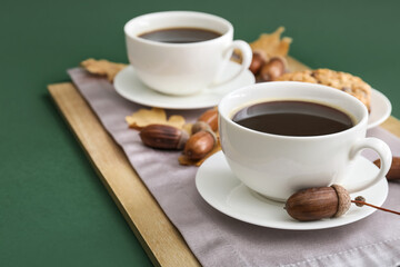 Board with cups of coffee with and acorns on green background, closeup