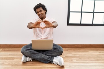 African man with curly hair using laptop sitting on the floor smiling in love doing heart symbol shape with hands. romantic concept.