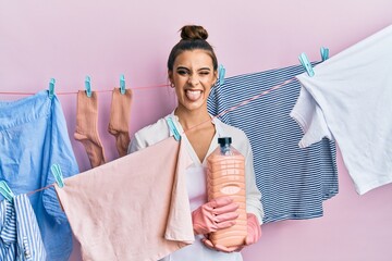 Beautiful brunette young woman doing laundry holding detergent bottle sticking tongue out happy...