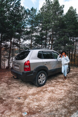 Woman tourist travel by SUV car for road trip in autumn. The traveler parking the car and enjoy beautiful scenery of forest in the background. Discovery and exploration. Vertical photo