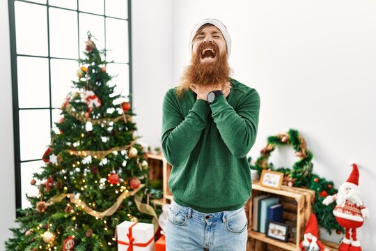 Redhead man with long beard wearing christmas hat by christmas tree shouting suffocate because painful strangle. health problem. asphyxiate and suicide concept.