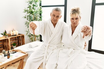 Middle age caucasian couple wearing bathrobe at wellness spa with angry face, negative sign showing dislike with thumbs down, rejection concept