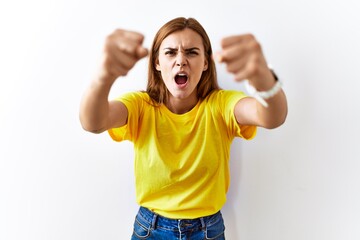 Young brunette woman standing over isolated background angry and mad raising fists frustrated and furious while shouting with anger. rage and aggressive concept.