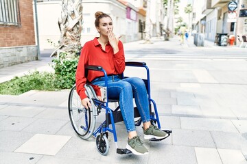 Young brunette woman sitting on wheelchair outdoors covering mouth with hand, shocked and afraid for mistake. surprised expression