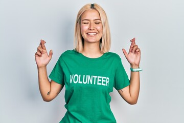Beautiful blonde woman wearing volunteer t shirt gesturing finger crossed smiling with hope and...