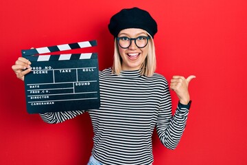 Beautiful blonde woman holding video film clapboard pointing thumb up to the side smiling happy...