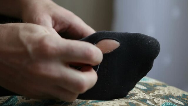 A man sews up black socks with white thread. The man mends his socks without taking them off.