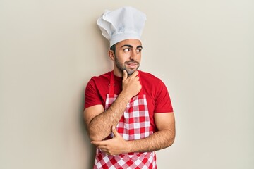 Young hispanic man wearing baker uniform with hand on chin thinking about question, pensive expression. smiling with thoughtful face. doubt concept.