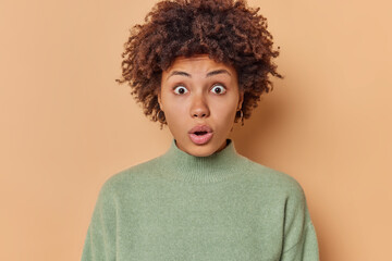 Portrait of stunned emotive beautiful woman with curly hair stares bugged eyes at camera looks...