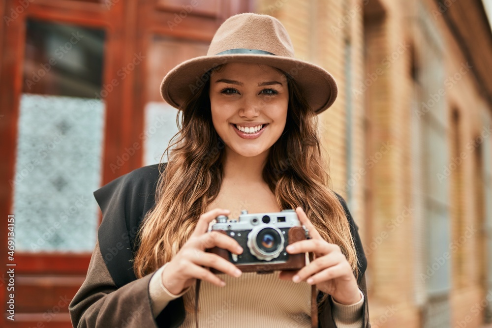 Wall mural Young hispanic tourist woman smiling happy using vintage camera at the city.