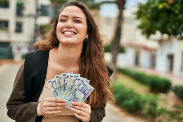 Young hispanic woman smiling happy holding romania leu banknotes at the city. - obrazy, fototapety, plakaty