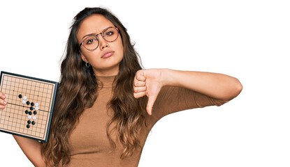 Young hispanic girl holding drauhgts with angry face, negative sign showing dislike with thumbs down, rejection concept