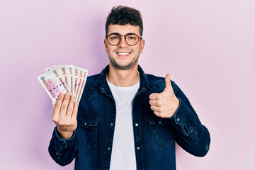 Young hispanic man holding saudi arabia riyal banknotes smiling happy and positive, thumb up doing excellent and approval sign