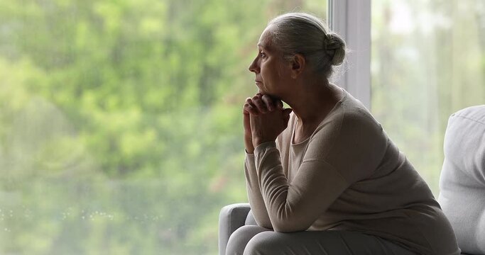 Side view of depressed older female sit on couch near picture window watch spring outside with sad look. Aged woman suffer of loneliness think of passed days of youth health problems brevity of life