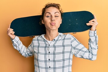 Young brunette woman holding skate puffing cheeks with funny face. mouth inflated with air,...