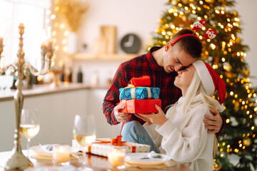 Cheerful young male and female Exchange of Christmas gifts. Young couple opening gift box near Christmas tree. Romantic day. Winter holidays.
