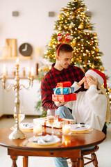 Cheerful young male and female Exchange of Christmas gifts. Young couple opening gift box near Christmas tree. Romantic day. Winter holidays.
