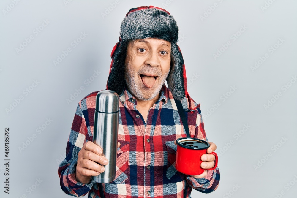 Sticker Handsome mature handyman wearing winter hat with ear flaps drinking hot coffee from thermo sticking tongue out happy with funny expression.